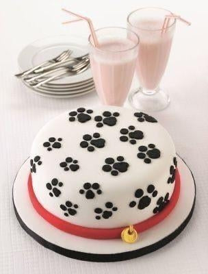 Pawprint celebration cake with black paw designs, a red ribbon, and pink milkshakes in the background.